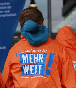 Zu sehen ist eine Person von hinten. Sie träge eine orange Jacke mit der Aufschrift "Journalismus ist MEHR WERT!"